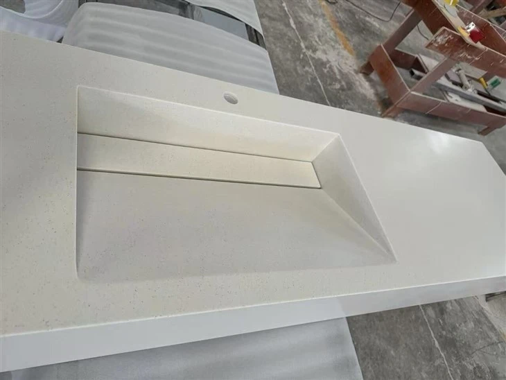 Close-up of a white acrylic integrated sink with sloped basin surfaces, linear drain cover, faucet hole and smooth finished joints.